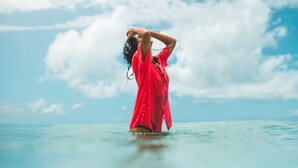 A person in a red shirt standing in clear turquoise water with a blue sky and clouds in the background. - Hotel Casa Coson (Las Terrenas)