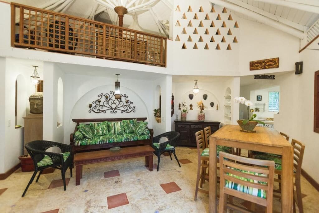 A spacious room with a high ceiling, a wooden dining table with chairs, a sofa with green cushions, and a decorative wall with triangular patterns.