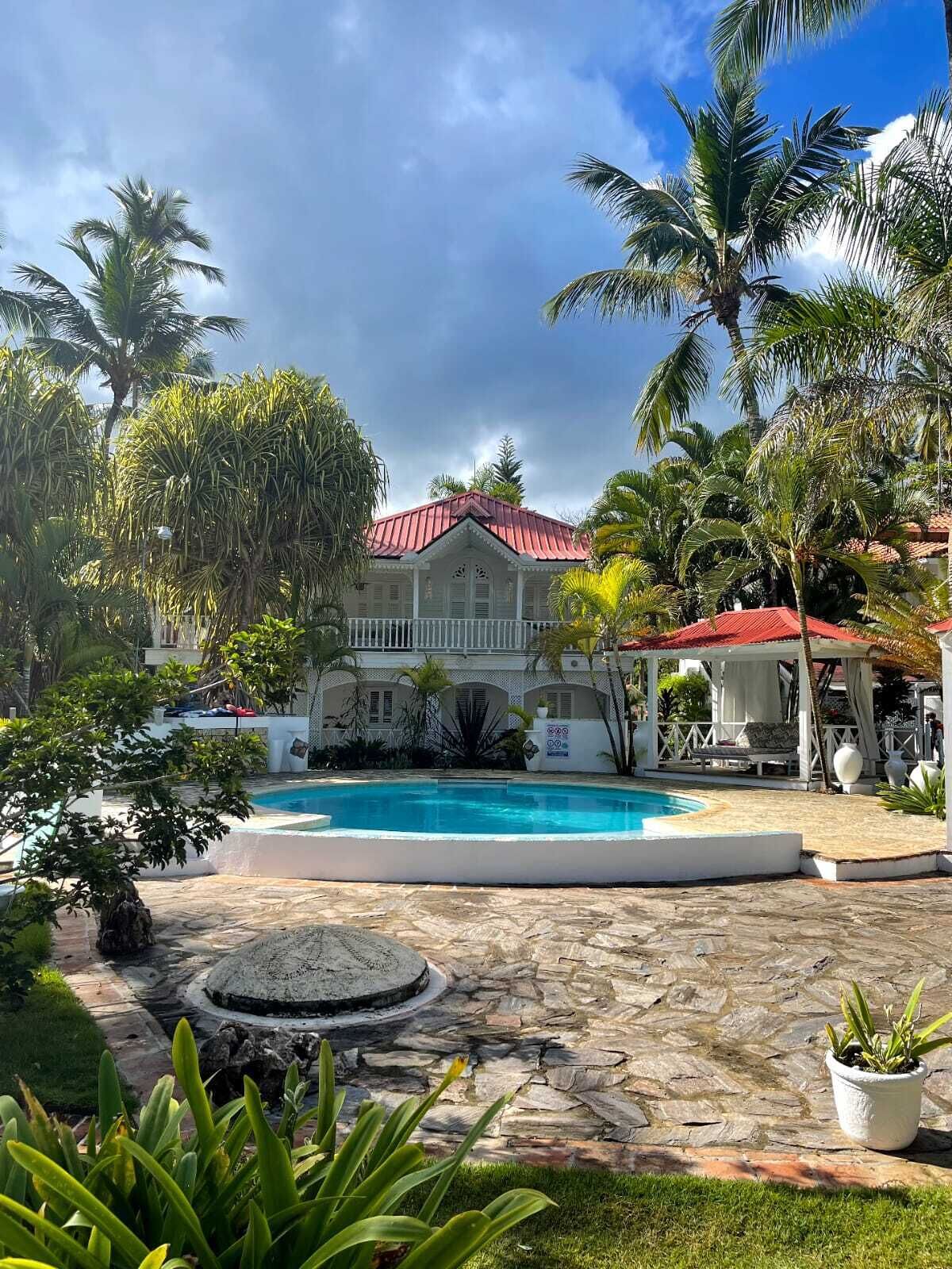 A pool area with a stone deck, a white building with a red roof, and palm trees.