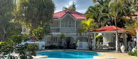 A pool area with a stone deck, a white building with a red roof, and palm trees.