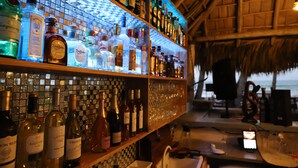 A bar with a variety of liquor bottles and glasses, a tiled backsplash, and a thatched roof. - Hotel Casa Coson (Las Terrenas)