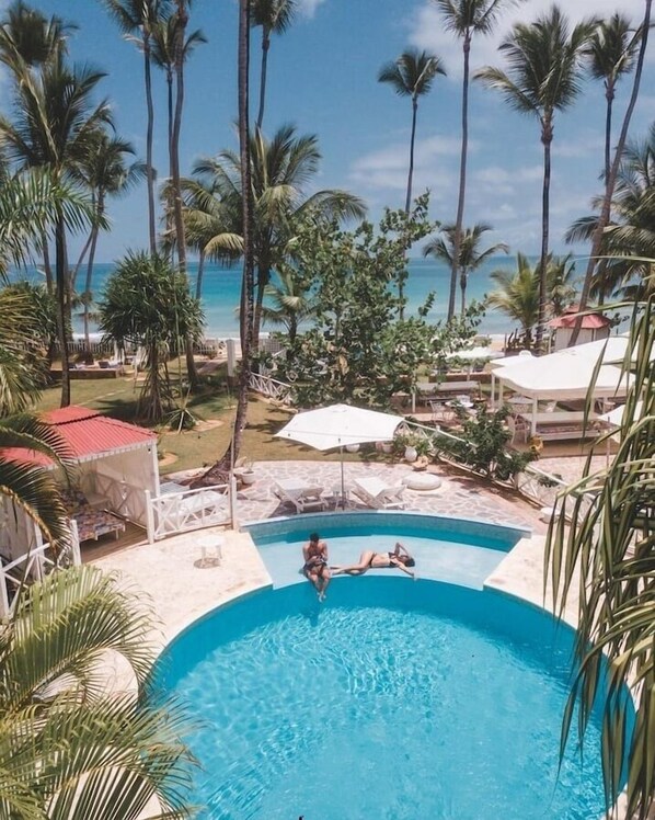 A swimming pool with two people sunbathing, surrounded by palm trees and a view of the ocean.