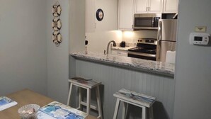 A compact kitchen with white cabinets, a microwave, a stove, and a built-in oven. There are two stools and a small table with magazines.