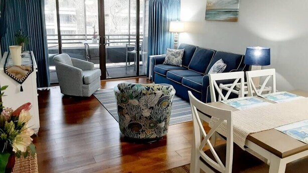 A living room with a blue sofa, a patterned armchair, a white table, and a view of the outdoors through large windows.