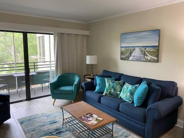 Un salon avec un canapé bleu foncé, un fauteuil turquoise et une table basse. Un tableau représentant une plage est accroché au mur, et la fenêtre offre une vue sur la verdure.