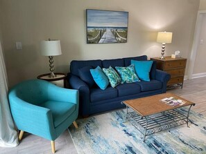 Een woonkamer met een donkerblauwe bank, een groenblauwe fauteuil en een salontafel met tijdschriften. Aan de muur hangt een schilderij van een strand en op een bijzettafeltje staat een lamp.
