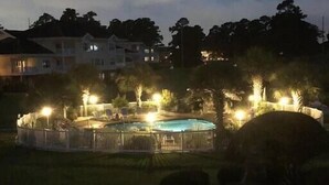 Área residencial com piscina cercada por grades, árvores e casas ao fundo.