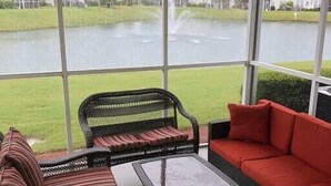 A covered patio with wicker furniture and a glass table, overlooking a lake and houses.