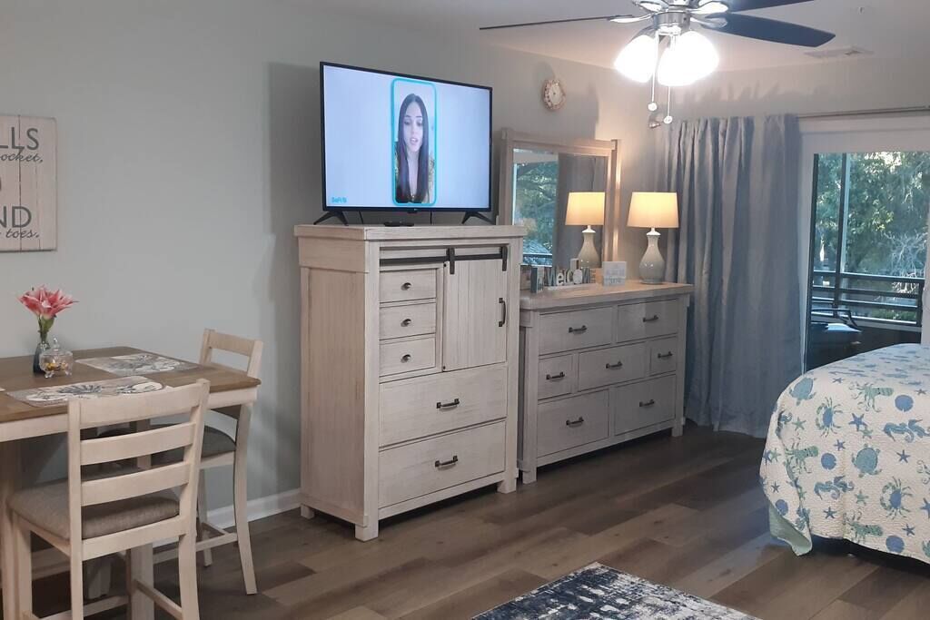 Ein Zimmer mit Holztisch, Fernseher, Kommode und einem Bett mit gemustertem Überwurf.