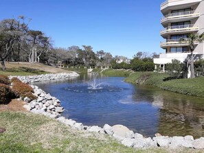 A tranquil pond with a fountain, surrounded by a grassy area and a multi-story building with balconies.