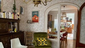 A living room with a green armchair, white chairs, a bookshelf, and a dining area visible through an open door. - Family home with large games room, close to the sea (Guilvinec)