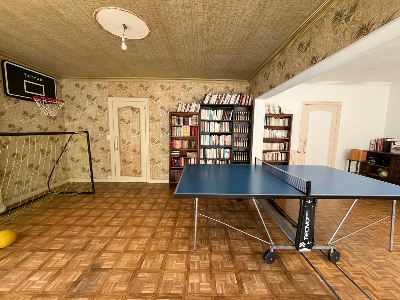 A room with a ping pong table, bookshelves, a basketball hoop, and a door.