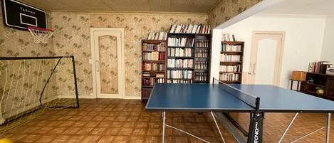 A room with a ping pong table, bookshelves, a basketball hoop, and a door.