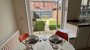 A dining area with a table set for four, a view of a garden and buildings outside, and a kitchen area visible to the right. - Kingmaker House - Luxury 3 Bedrooms, Driveway, Garage, Garden, M1 (Northampton)