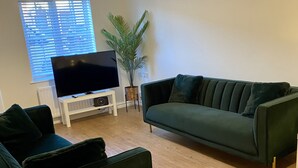 A living room with a dark green sofa, a white entertainment unit with a flat-screen TV, a potted plant, and a window with blinds. - Kingmaker House - Luxury 3 Bedrooms, Driveway, Garage, Garden, M1 (Northampton)