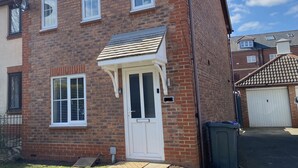 A two-story brick house with a white front door, a small porch, and a garage door. - Kingmaker House - Luxury 3 Bedrooms, Driveway, Garage, Garden, M1 (Northampton)