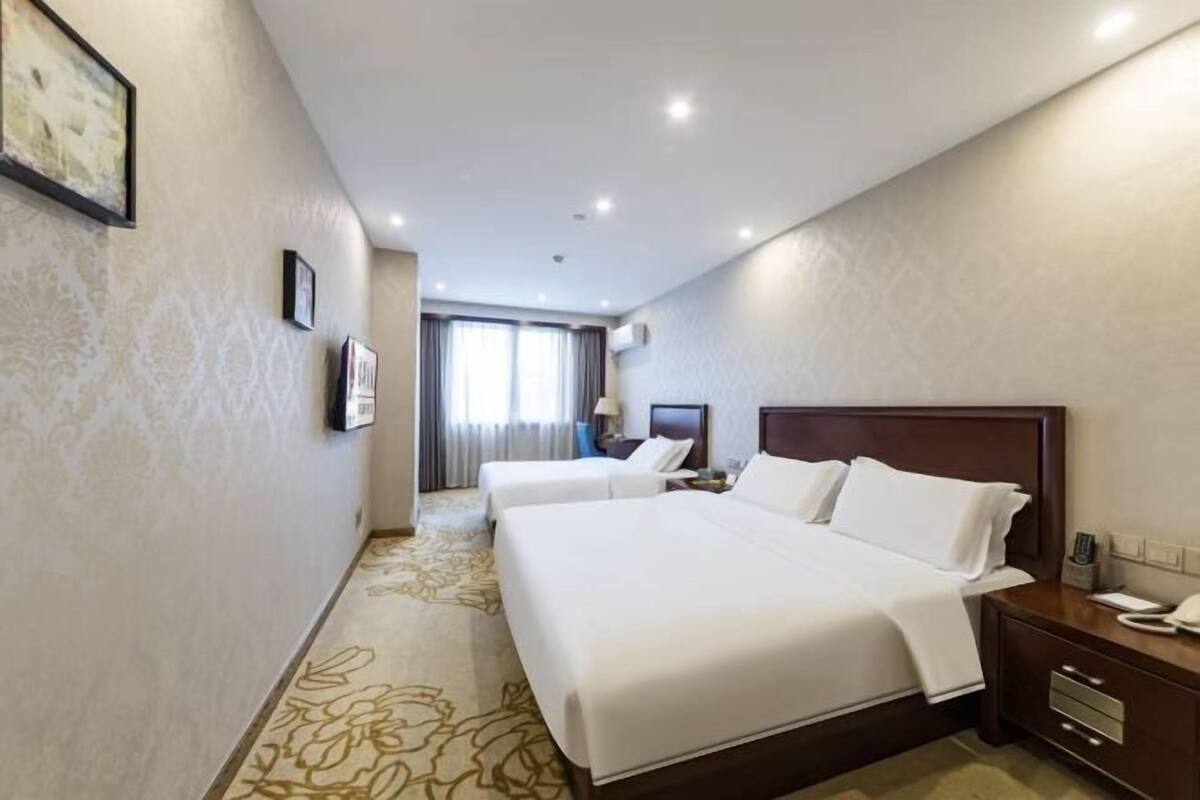 A hotel room with two beds, a wooden headboard, a bedside table, a nightstand, and a large window.