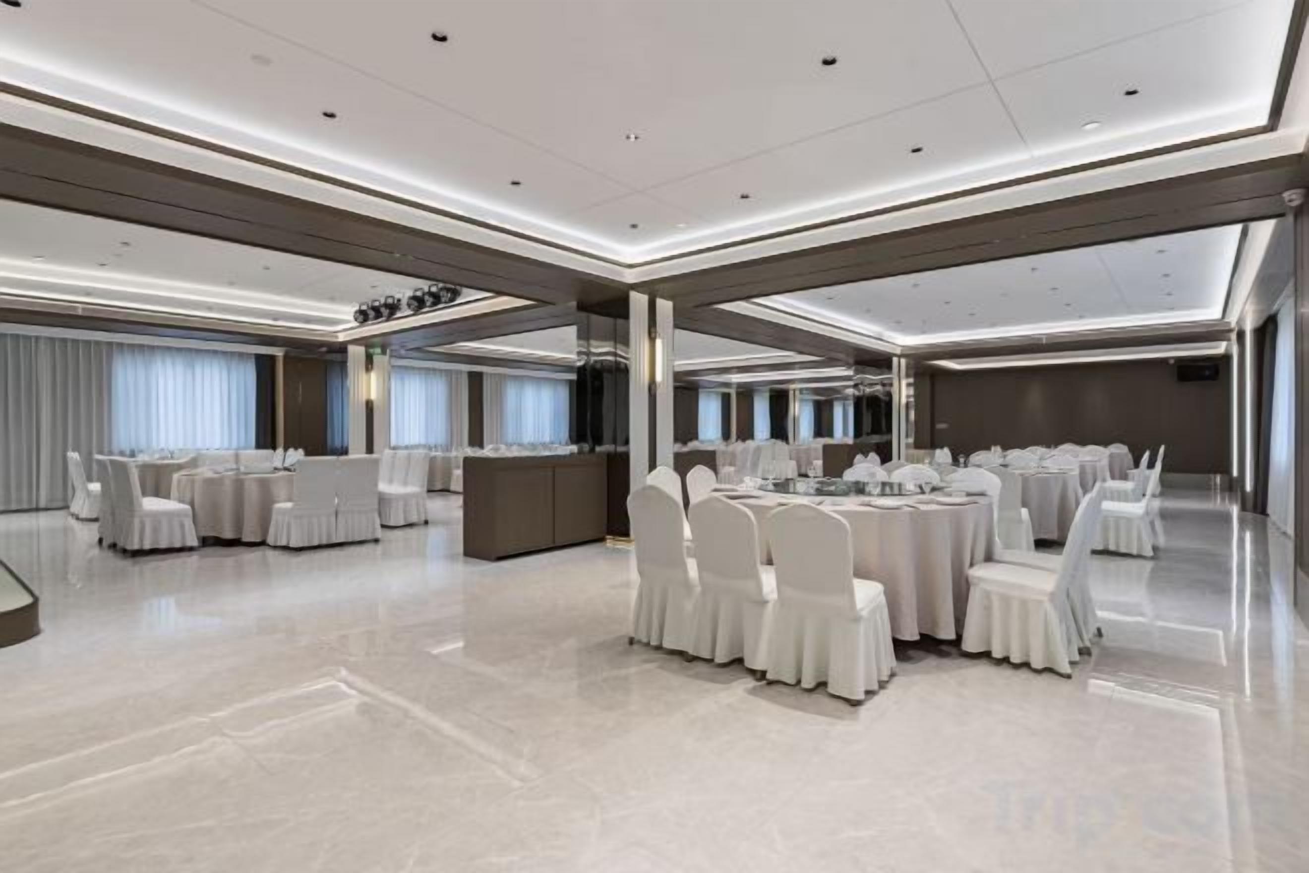 Un amplio salón de banquetes con mesas redondas y sillas blancas, una zona de recepción y un diseño de techo moderno.
