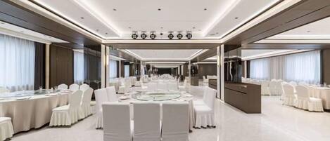 Un amplio salón de banquetes con mesas y sillas redondas, una zona de bar central y un largo pasillo con ventanas.