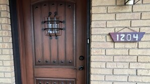 Una porta de fusta marró amb una aldreba de metall i un petit graó que hi mena. Es veu una paret de rajola amb un senyal numèric '1204' a la dreta de la porta.