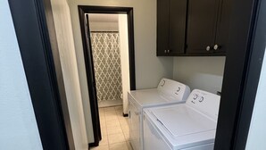 Una sala de bugaderia amb dues rentadores blanques, una llum al sostre i una porta que condueix a una altra habitació.