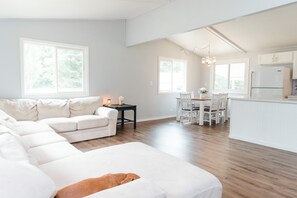 Amplia sala de estar con un sofá seccional blanco grande, un comedor con mesa y sillas de madera, y una cocina con gabinetes blancos y un refrigerador.