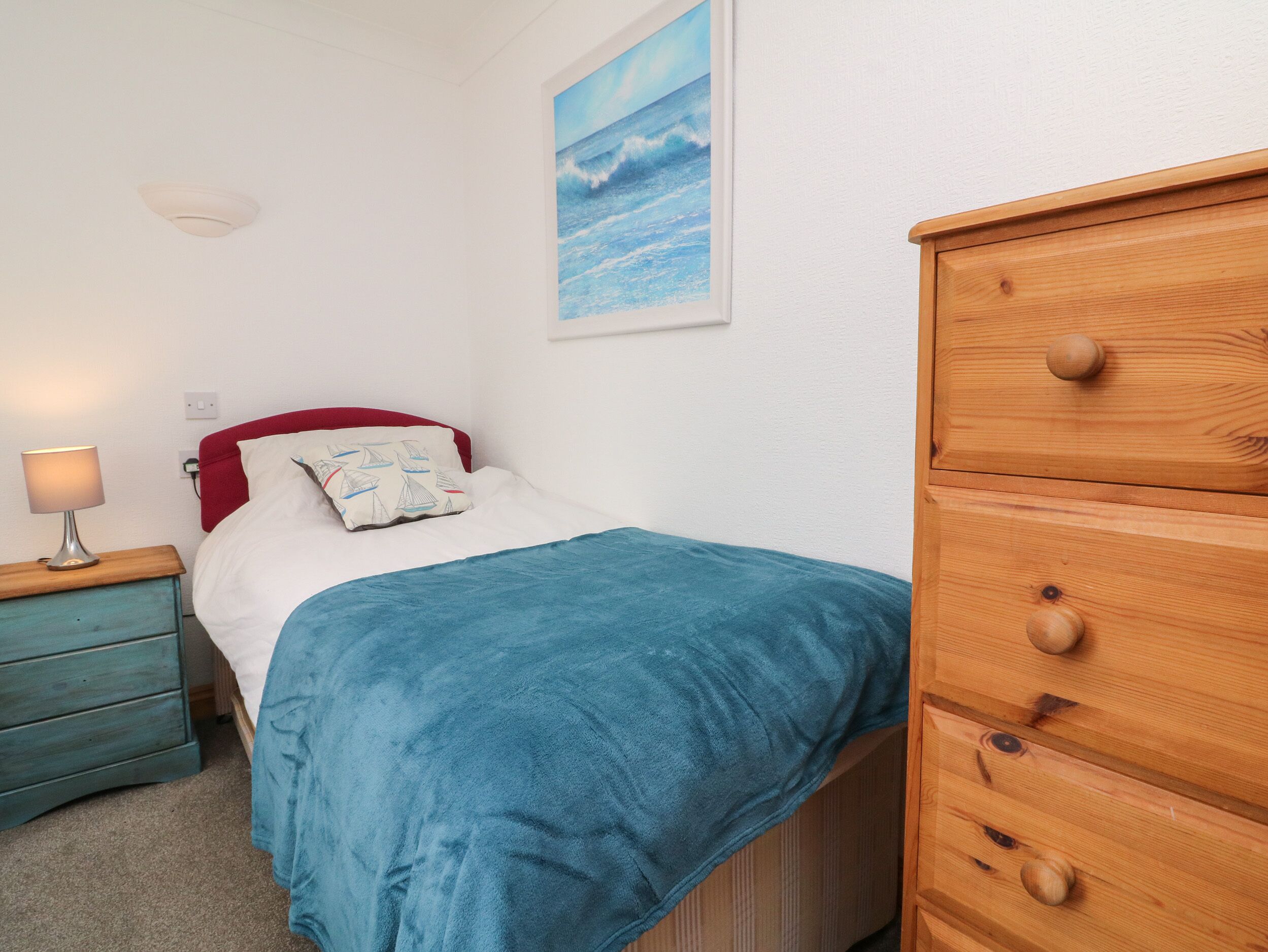 A bedroom with a bed, a wooden dresser, a nightstand with a lamp, and a painting on the wall.