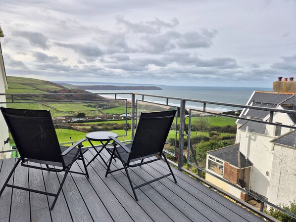 A balcony with two chairs and a table, overlooking a coastal landscape with hills and a beach. - 5 Seymour Villas (Woolacombe)