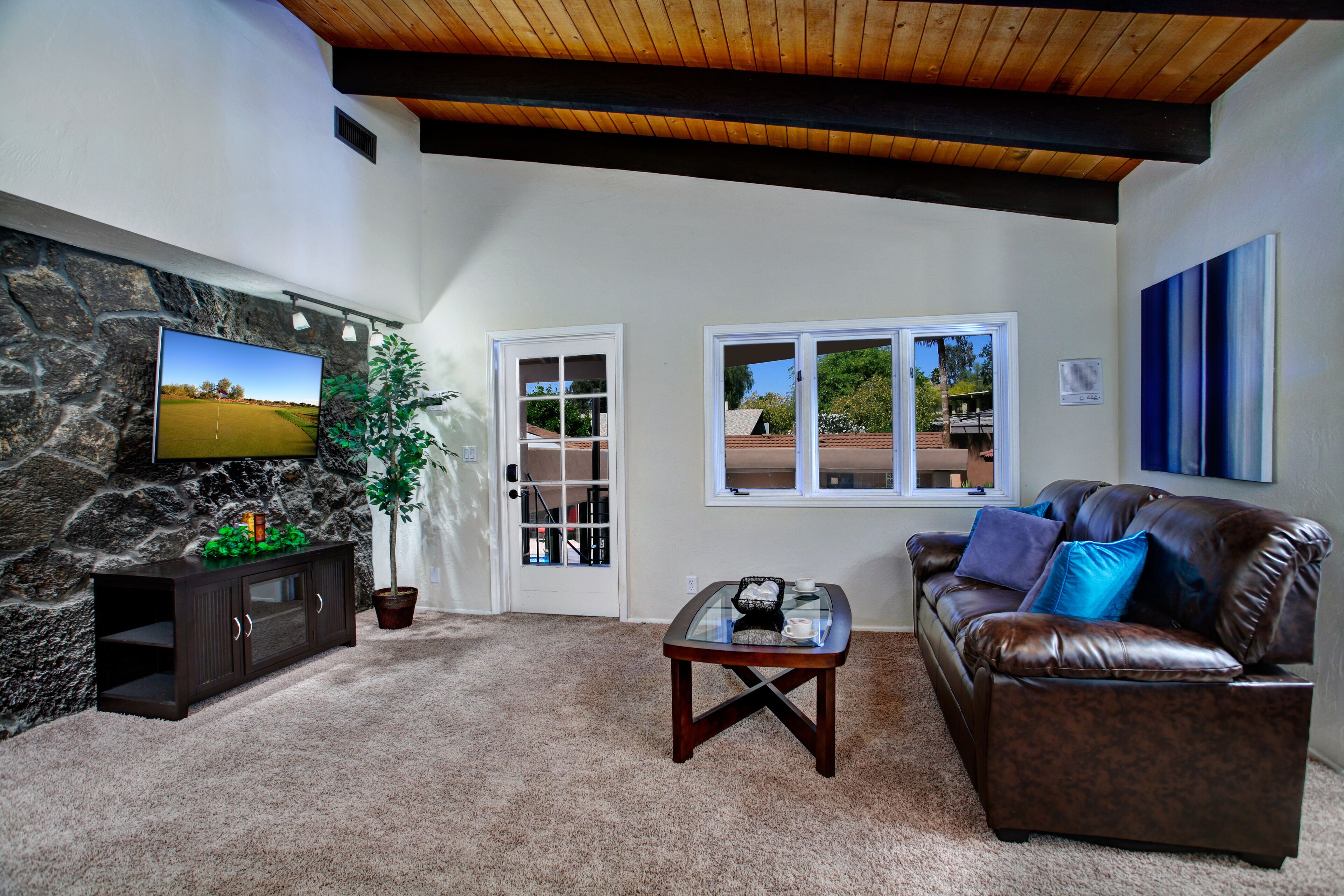 A living room with a stone wall, a television, a leather couch, a glass coffee table, and a door leading outside.