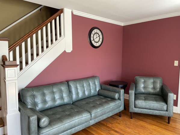 A living room with a grey leather sofa, a matching armchair, a round side table, and a wooden staircase.