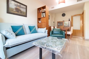 A living room with a sofa, a glass coffee table, a wooden bookshelf, a dining table with chairs, and a clock on the wall.