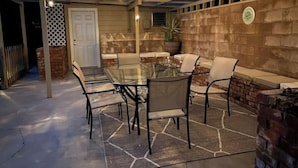 Un patio al aire libre con una mesa y sillas de vidrio, un muro de ladrillo y una pérgola con luces colgantes.