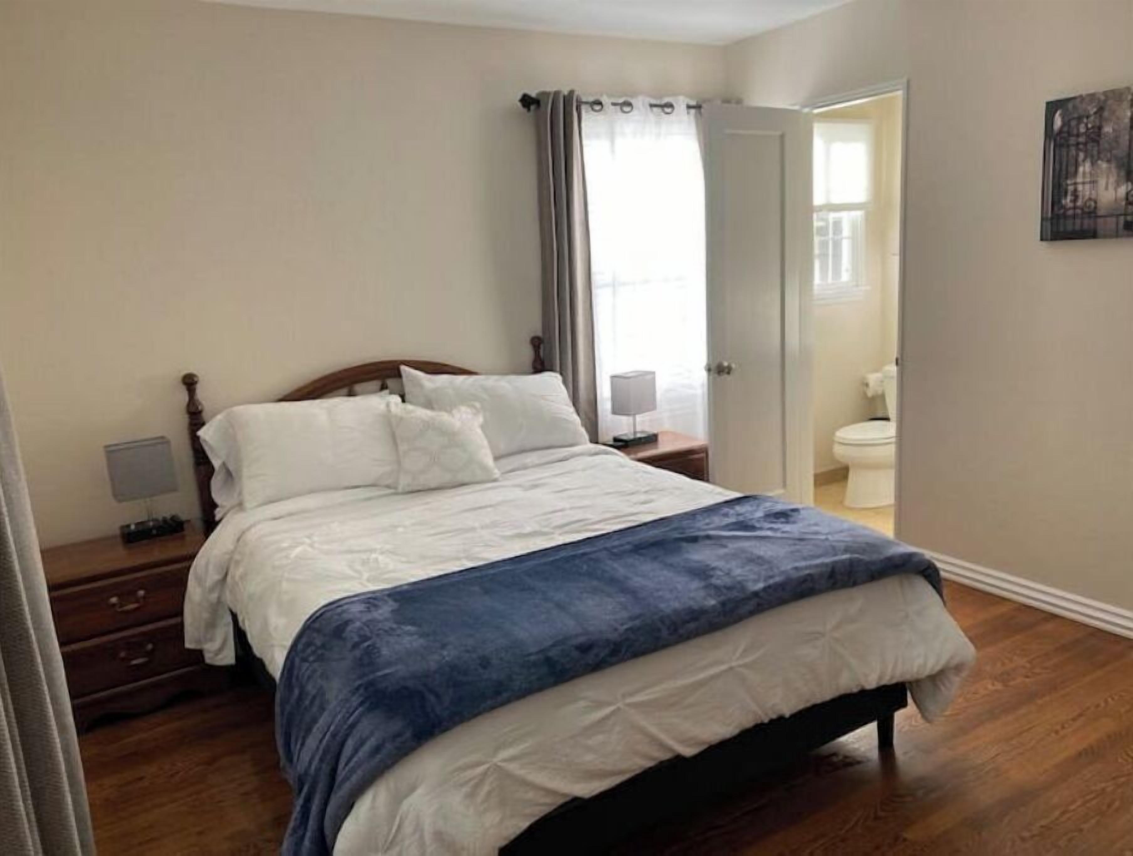A bedroom with a bed, a nightstand, a drawer, a lamp, and a bathroom visible through an open door.
