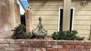 Una estatua de meditación sobre un borde de ladrillo, con un muro de ladrillo y una puerta de madera al fondo.