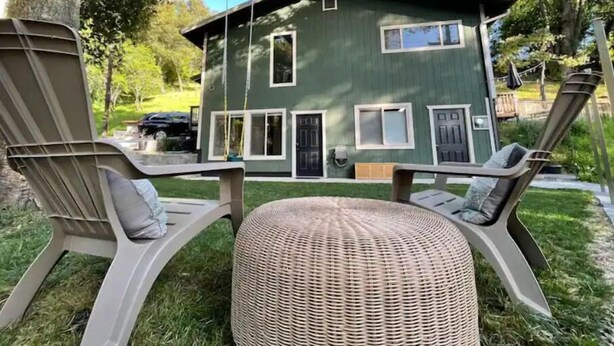 A two-story house with a green exterior, a front door, and a small porch area with two chairs and a wicker ottoman.