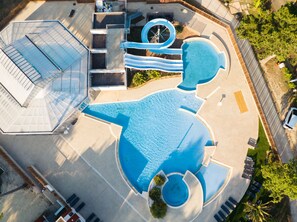 Vista aérea de un parque acuático con un tobogán azul grande, una piscina y una zona cubierta con asientos.