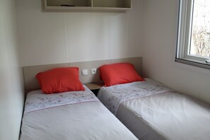 Una habitación pequeña y limpia con dos camas individuales, cada una con almohada roja y ropa de cama floral. Se ve una ventana a la derecha y una estantería blanca encima de las camas.