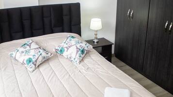 A neatly made bed with a quilted headboard, two decorative pillows, and a folded towel. A small bedside table with a lamp is visible.