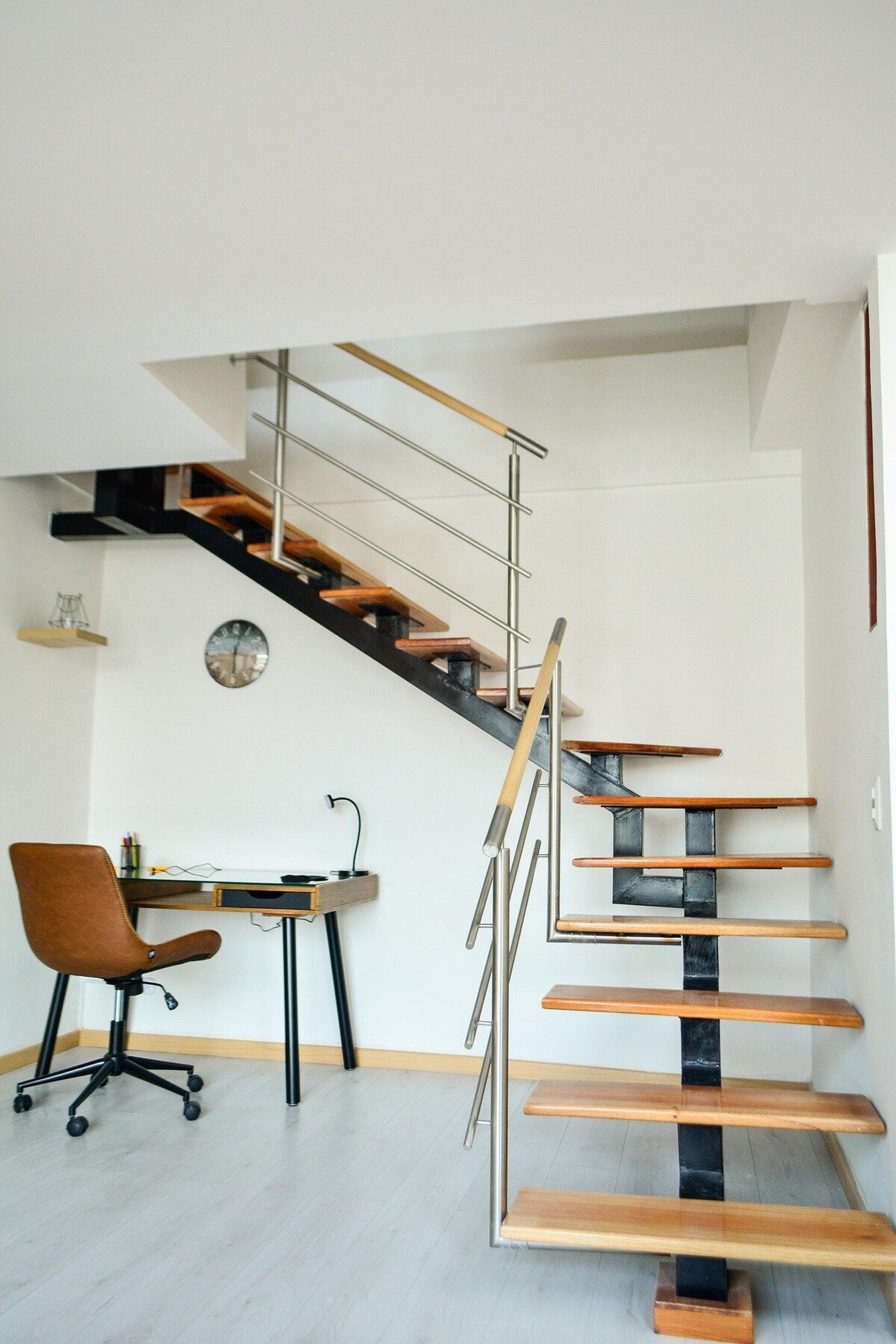 A modern staircase with wooden steps and metal railings, a desk with a chair, and a clock on the wall.