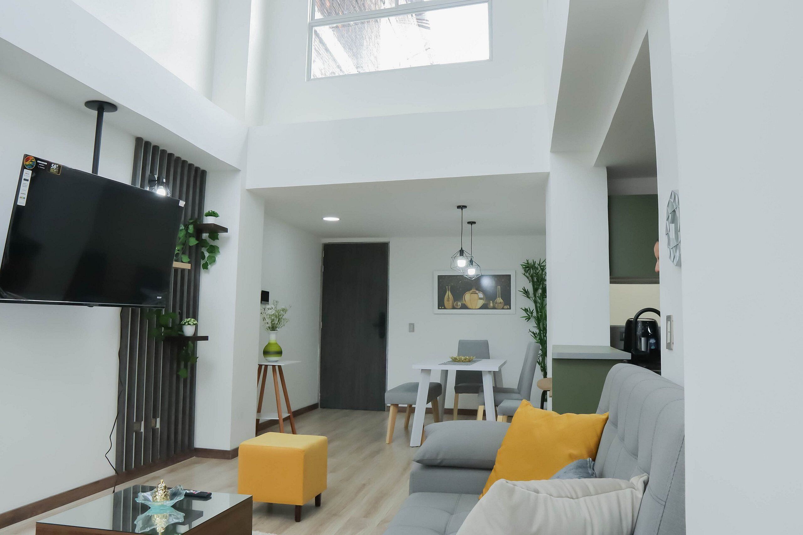 Un salon moderne avec un téléviseur à écran plat, une table basse en verre, un canapé avec des coussins, un coin repas avec une table et des chaises, et un espace cuisine en arrière-plan.