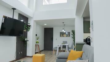 Un salon moderne avec un téléviseur à écran plat, une table basse en verre, un canapé avec des coussins, un coin repas avec une table et des chaises, et un espace cuisine en arrière-plan.
