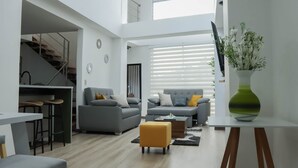 A modern living room with two grey sofas, a yellow ottoman, a coffee table, and a large window with blinds. - EDIFICIO CAMILA (Manizales)