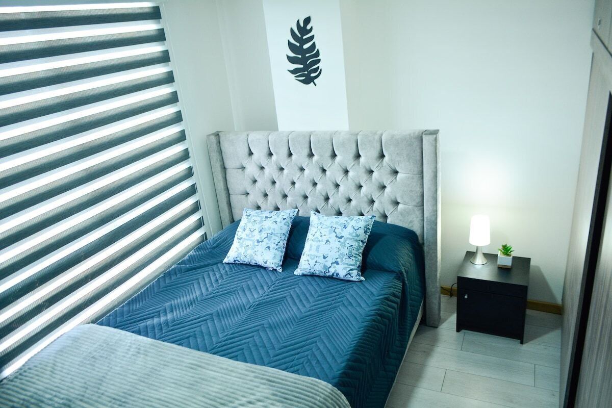 A bedroom with a tufted headboard, a bed with a blue quilt, two matching pillows, a nightstand with a lamp, and a wall-mounted leaf decoration.
