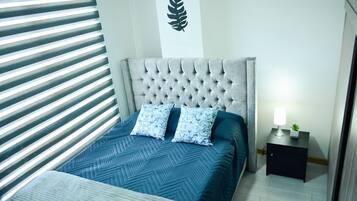 A bedroom with a tufted headboard, a bed with a blue quilt, two matching pillows, a nightstand with a lamp, and a wall-mounted leaf decoration.