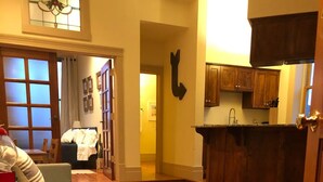 A room with wooden flooring, a white table, a grey sofa, and a red object on the table. There is a doorway leading to another room with a guitar on the wall.