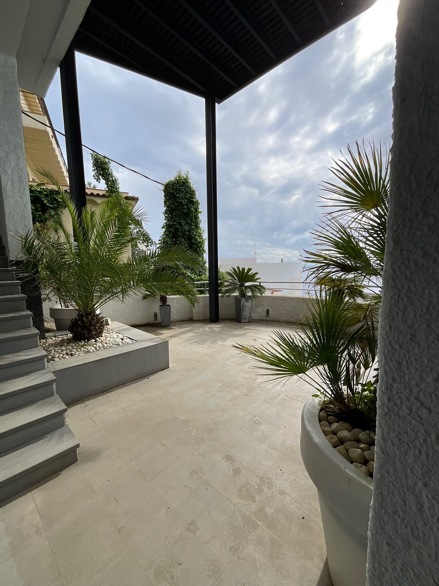 A covered outdoor area with a concrete floor, surrounded by potted plants and a staircase leading to an upper level.