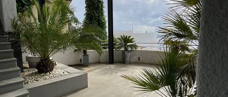 A covered outdoor area with a concrete floor, surrounded by potted plants and a staircase leading to an upper level.