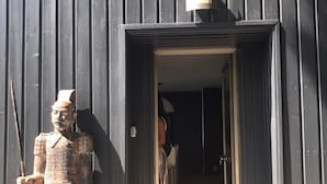 Une porte en bois ornée d’une statue représentant un guerrier tenant une lance et un bouclier, un petit luminaire placé au-dessus de la porte, et un étroit couloir menant à l’intérieur.