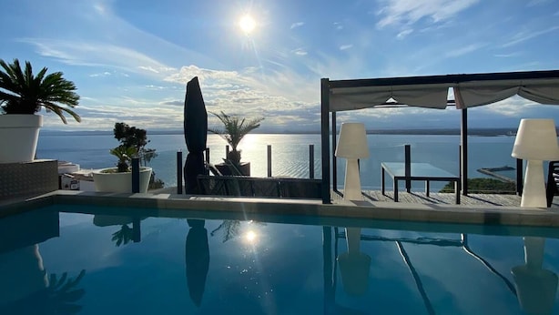 Un ciel d’un bleu éclatant, avec le soleil qui brille sur un espace piscine agrémenté de chaises longues et de plantes en pot.
