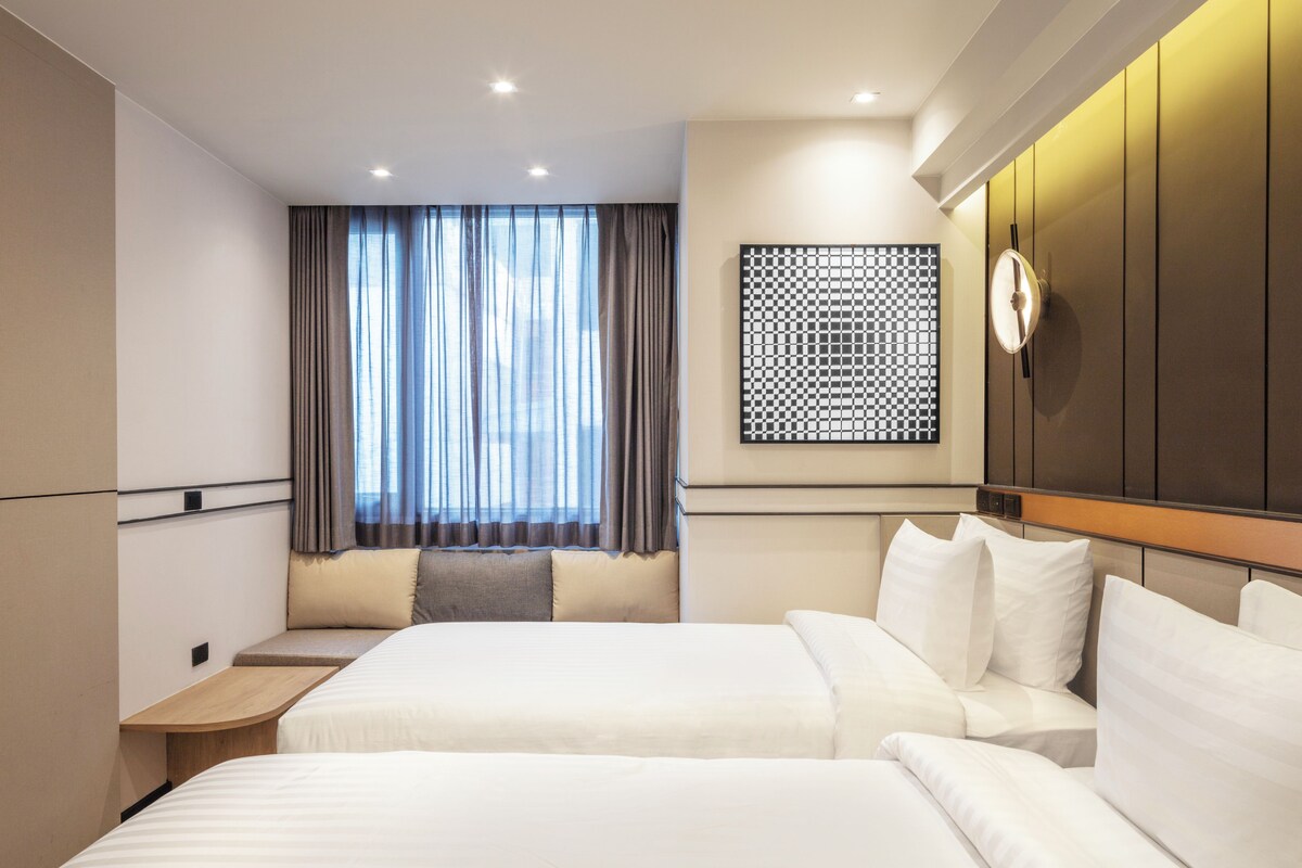 A modern hotel room with two beds, a large window with curtains, a wall-mounted clock, and a decorative panel.